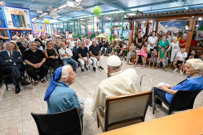 A nun in blue and a religious leader speak to an attentive audience in a colorful meeting room.