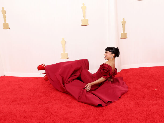 Celebrity in a red gown, sitting playfully on the red carpet, showcasing fashion challenges.