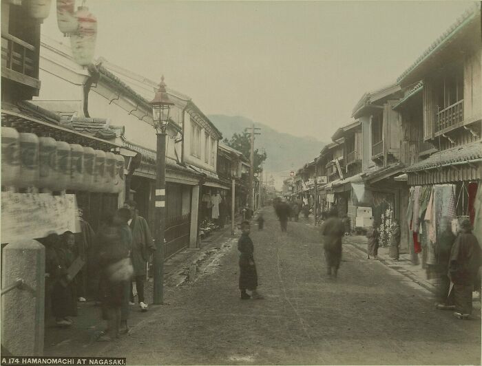 Street scene in 1800s Japan with traditional wooden buildings and people walking along a dirt road at Nagasaki.