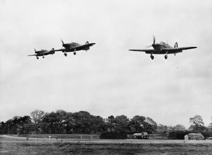 Three WWII fighter planes flying in formation over a landscape with trees and a vehicle below.