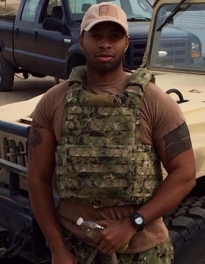 Former Navy SEAL in tactical gear holding a bottle, standing in front of a vehicle.