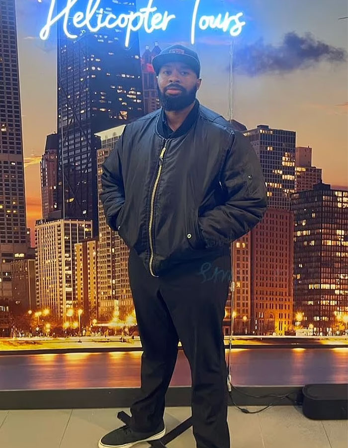 Man in front of cityscape backdrop under "Helicopter Tours" sign, wearing a black jacket and cap.
