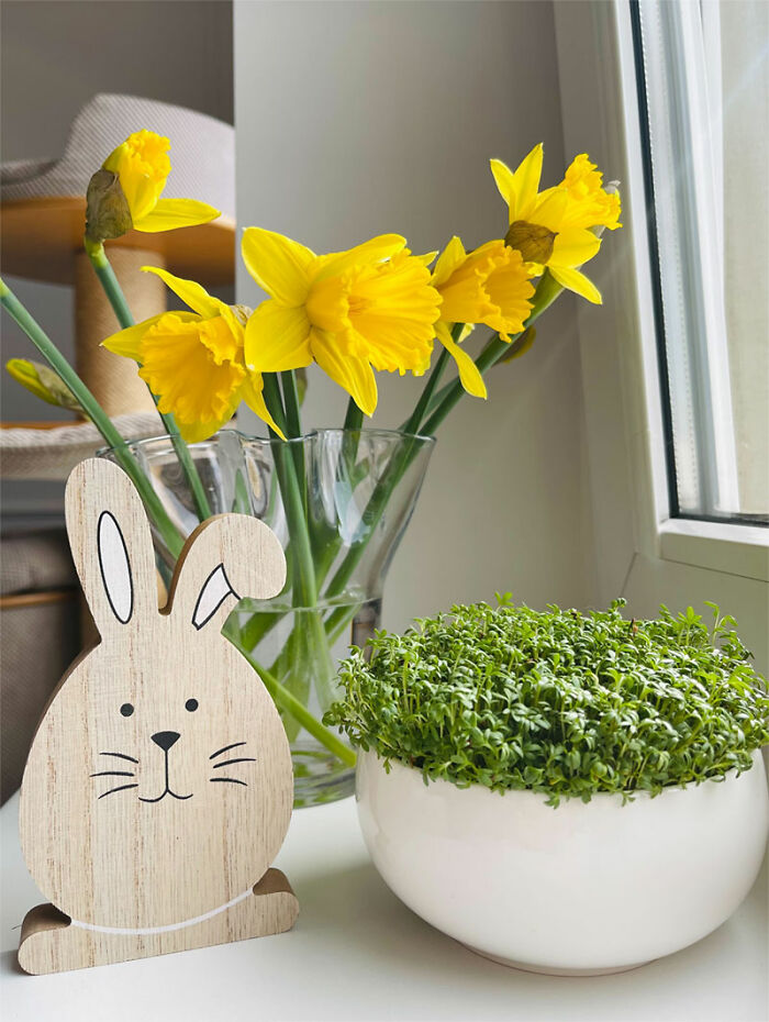 Easter decorations featuring a wooden bunny, yellow daffodils in a glass vase, and greenery in a white bowl on a windowsill.