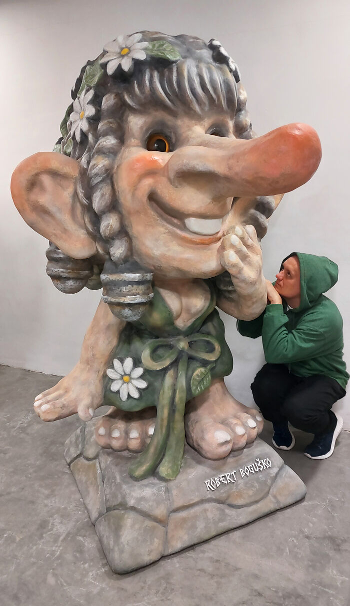 Whimsical troll sculpture with daisy decorations, crafted from outdoor materials, person in a green hoodie nearby.