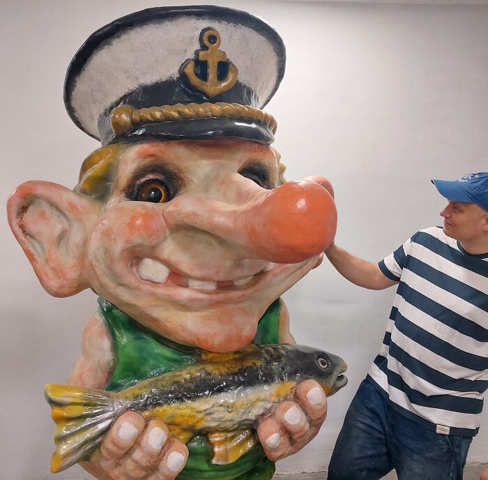 Man standing beside large whimsical troll sculpture holding a fish, wearing a sailor hat.