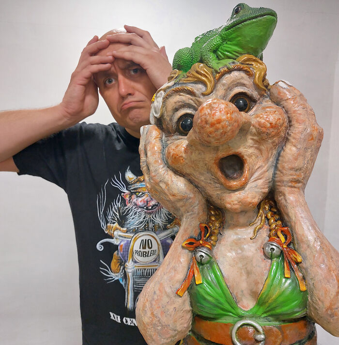 Man with whimsical troll sculpture featuring a frog on its head, expressing surprise.