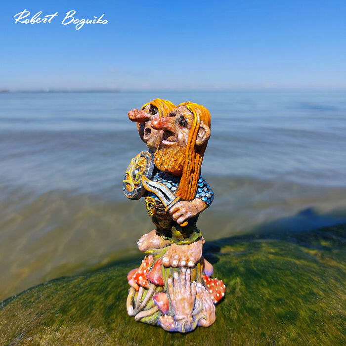 Whimsical troll sculpture made from outdoor materials, standing by the sea under a clear blue sky.