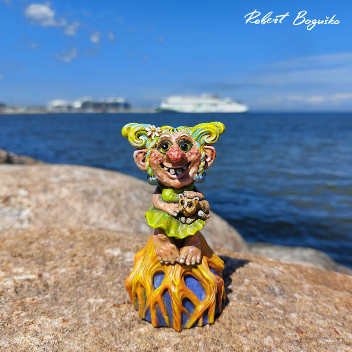 Whimsical troll sculpture with colorful hair, sitting on rocks by the sea, cruise ship in the background.