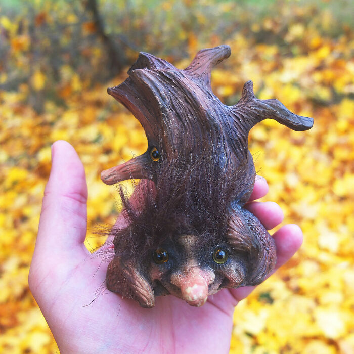 Hand holding a whimsical outdoor troll sculpture with earthy textures and vibrant fall leaves in the background.