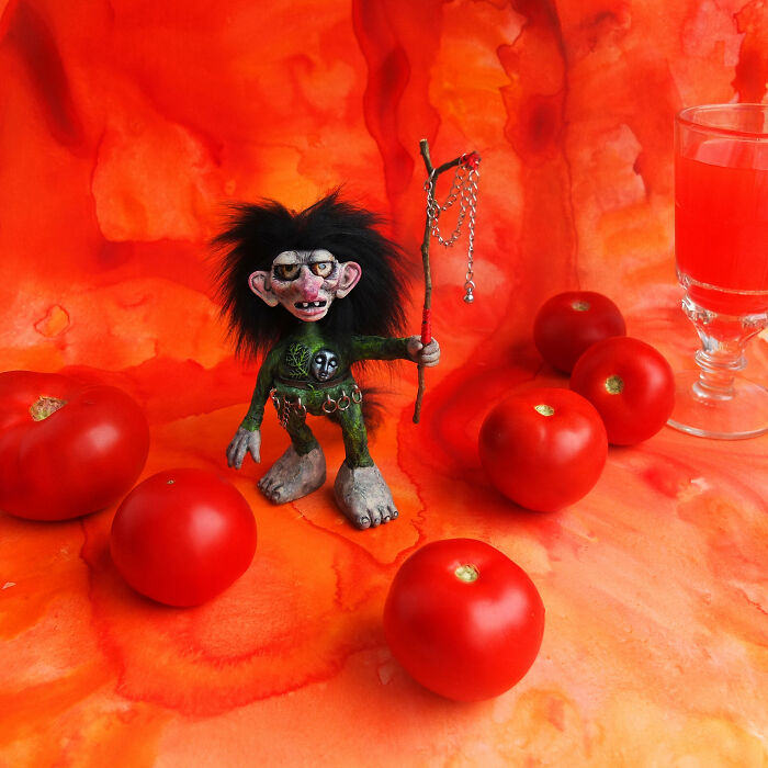 Whimsical troll figurine amidst tomatoes and a drink, set against a vibrant orange background.