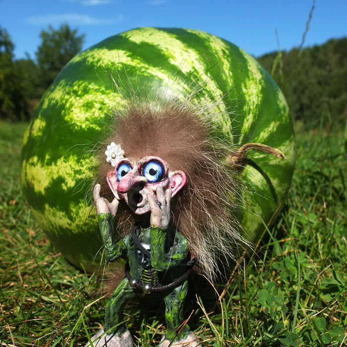 Whimsical troll with large eyes in front of a watermelon, made from outdoor materials, on grass.