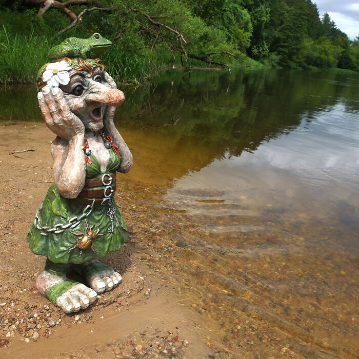 Whimsical troll sculpture by a riverside, adorned with chains and a frog, created from outdoor materials.