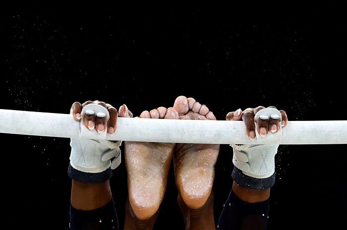 Gymnast grips the uneven bars, chalk on hands and feet, in a dynamic Istanbul Photo Awards 2025 image.