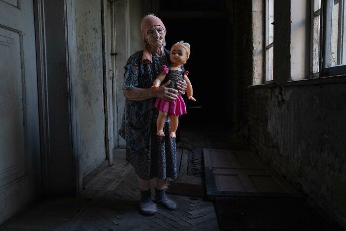 Elderly woman holding a doll in a dimly lit hallway, featured in Istanbul Photo Awards 2025.