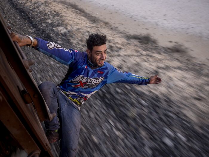 Man in a blue racing shirt joyfully hangs off a moving train, showcasing powerful imagery from Istanbul Photo Awards 2025.