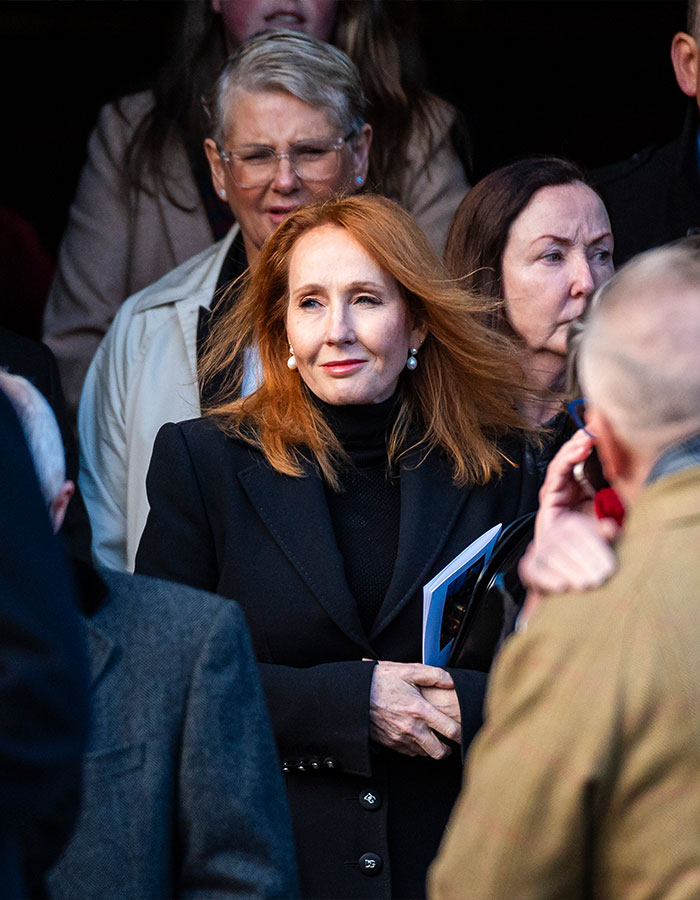 A woman with red hair in a group, wearing a black coat, holding a booklet.