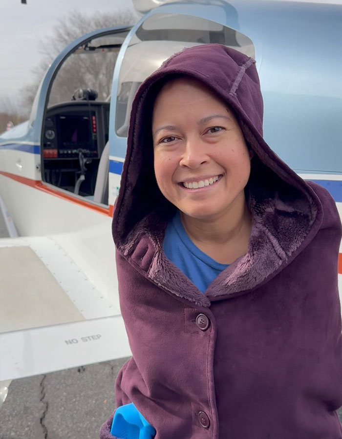 Person with a smile, wearing a hooded coat, standing by a small airplane. Real-life superpowers theme.