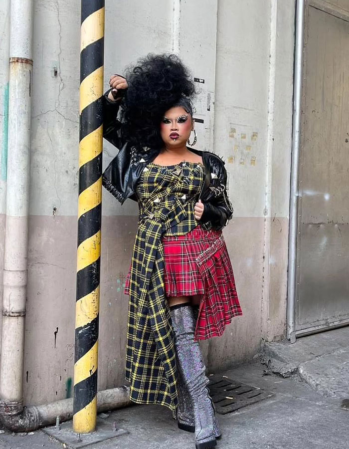 Drag Race alum in bold outfit with plaid patterns and metallic boots, standing by a striped pole outdoors.
