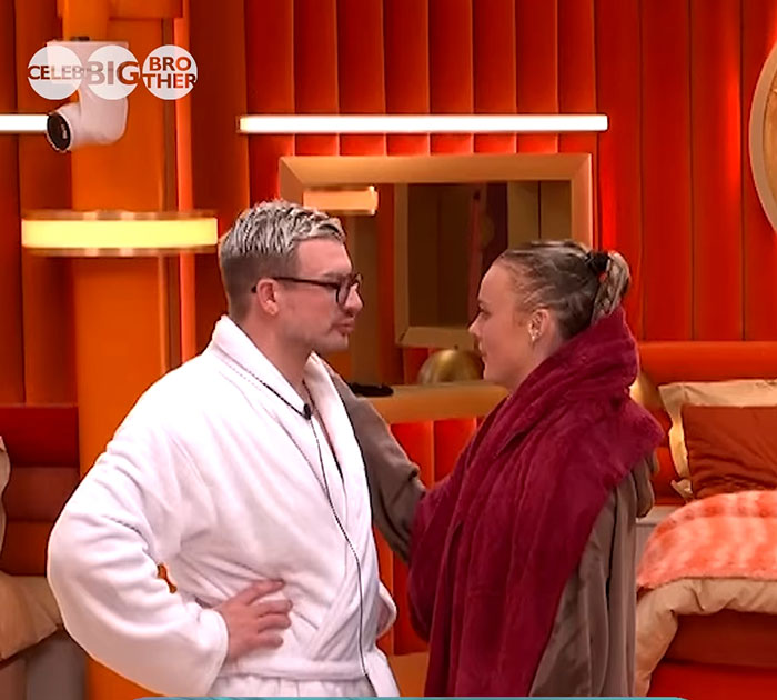 Two contestants share a moment in the Big Brother house, standing in a bright, orange-themed room.