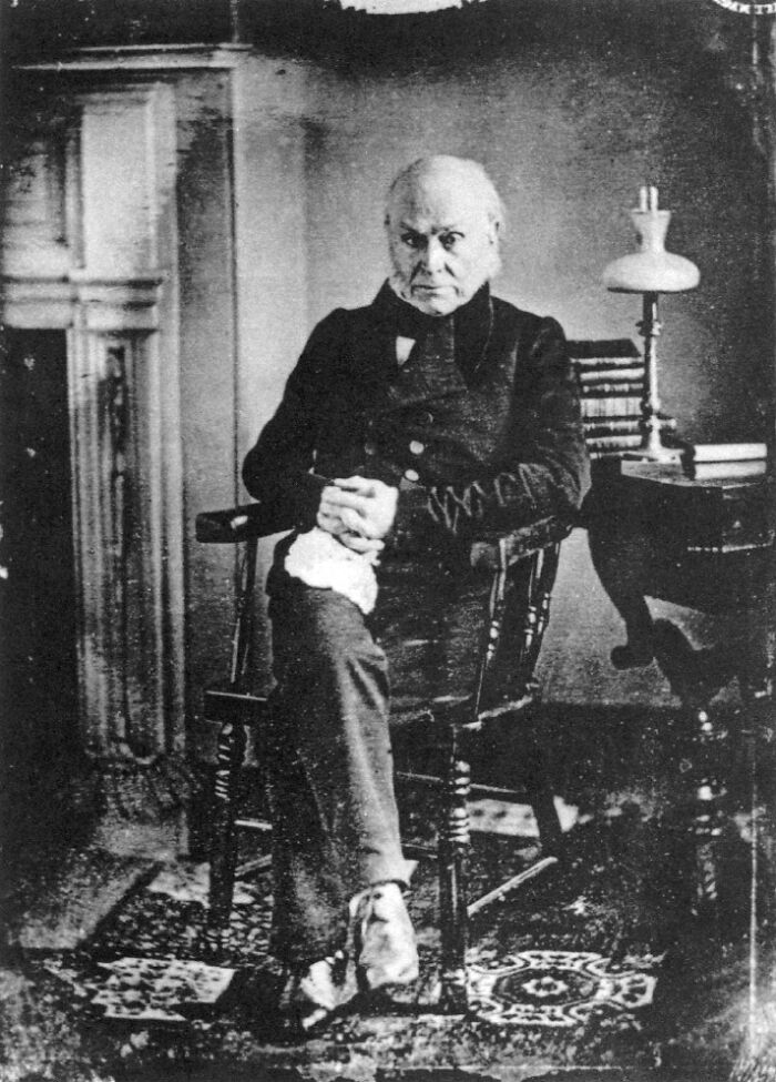 Rare photo of a famous historical figure seated in a chair inside a room with vintage furniture and books.