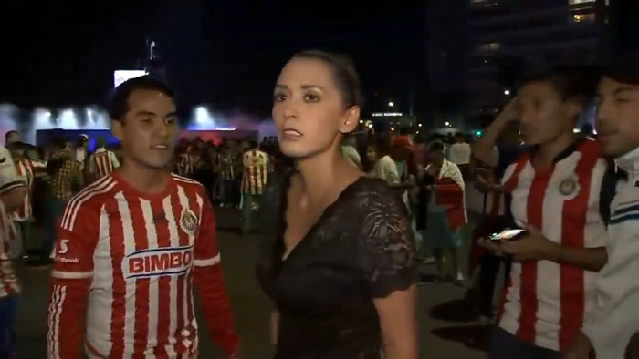 A female reporter looks concerned while surrounded by fans in sports jerseys at a nighttime event.