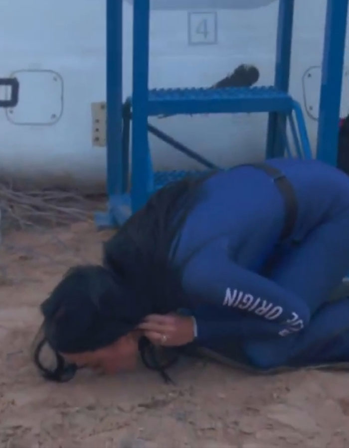 Person in blue space suit kissing the ground, highlighting return from space trip.