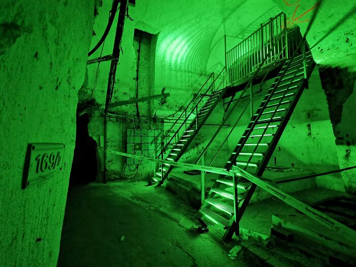Eerie underground city scene with green lighting, showing metal stairs and worn walls.