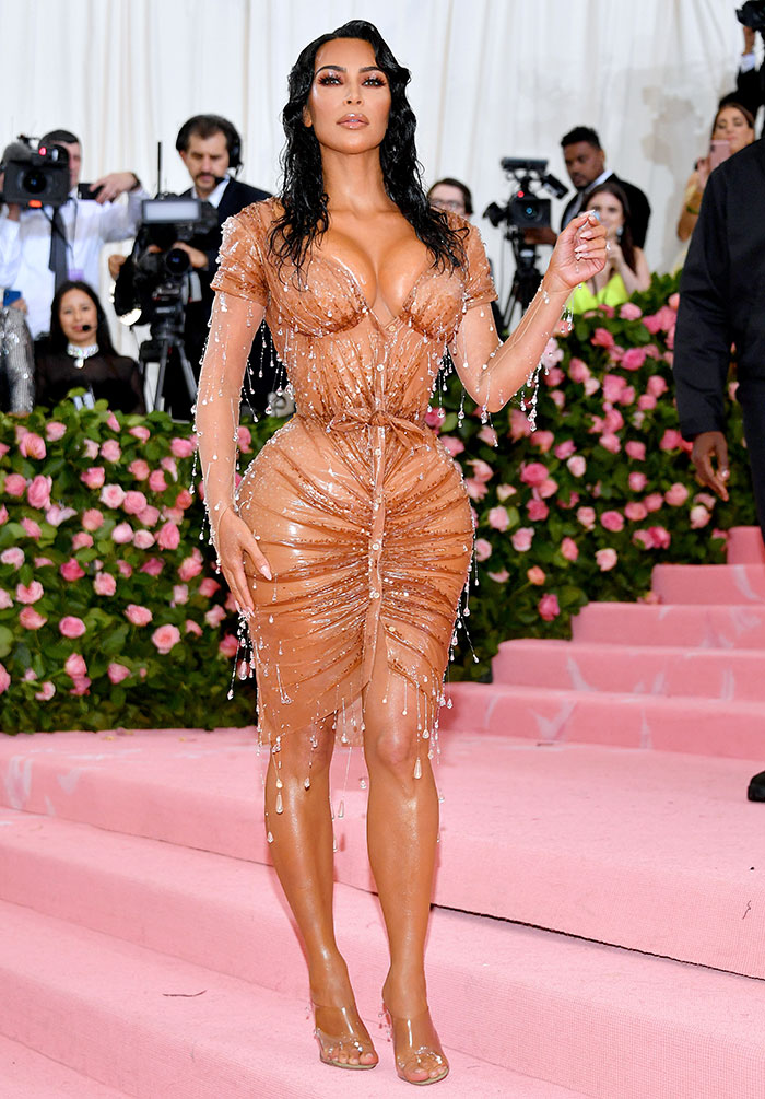 Woman in elegant wet-look dress on red carpet with cameras in background, showcasing glamorous fashion amid behind-the-scenes moments.