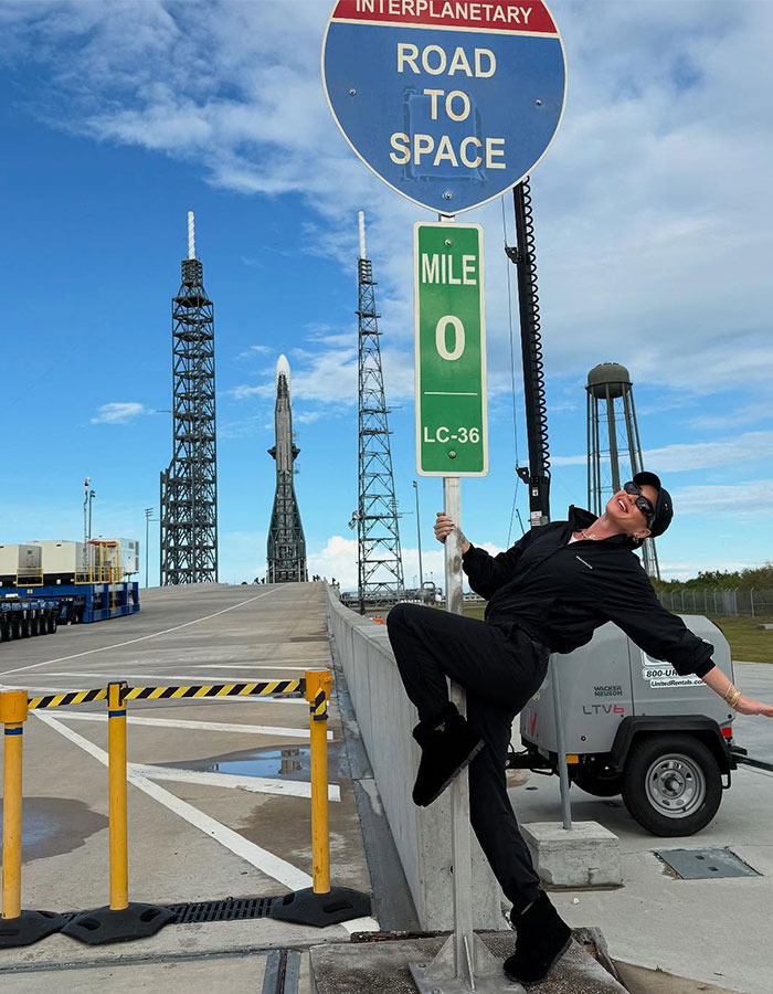 Person in black outfit posing under "Road to Space" sign at a space launch site, highlighting space mission theme.