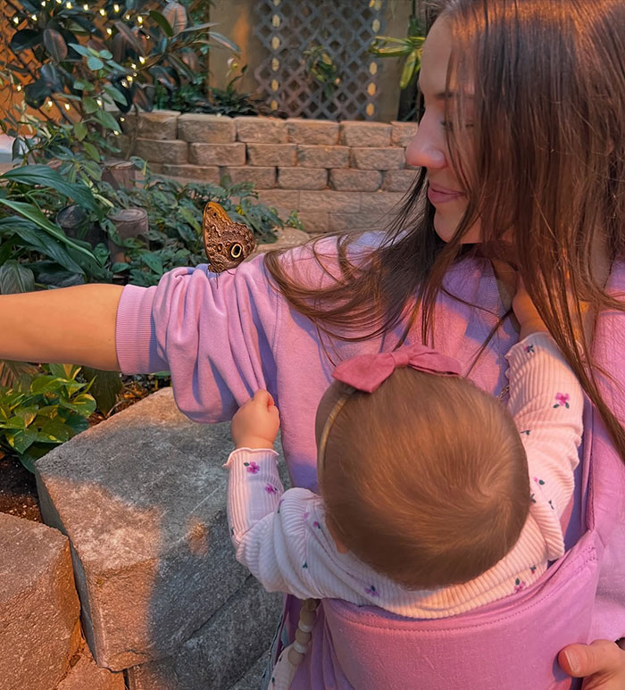 Influencer holding her baby, both observing a butterfly perched on her arm in a garden setting.