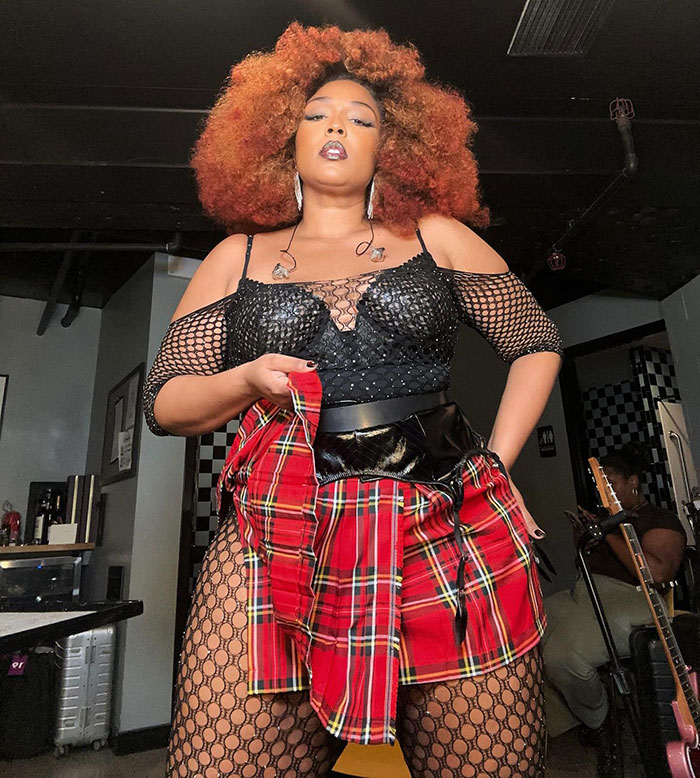 Lizzo in a bold outfit, posing confidently backstage with a checkered skirt and black mesh top.