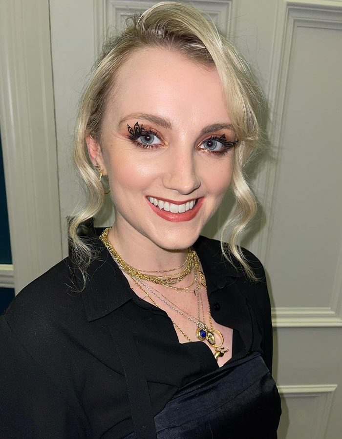 Harry Potter star smiling in elegant attire, includes necklace and unique eye makeup, standing against a wooden panel. Harry Potter star smiling in elegant attire, includes necklace and unique eye makeup, standing against a wooden panel.