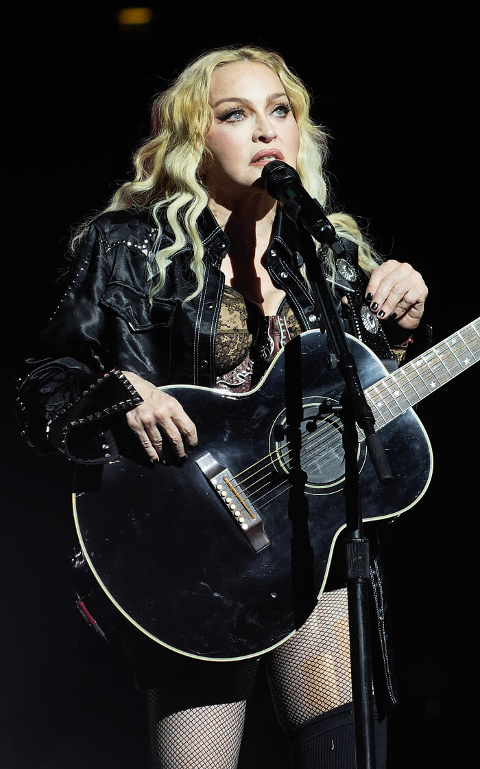 Celebrity performing live with a guitar, wearing a leather jacket and fishnet stockings.