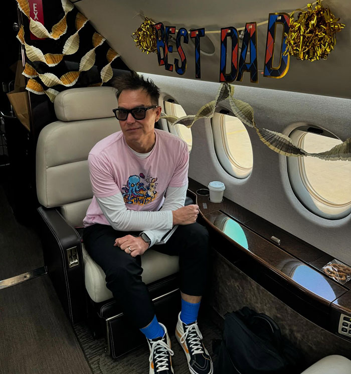Mark Hoppus from Blink-182 sits in a private jet with "Best Dad" banner, wearing a pink shirt and sunglasses.
