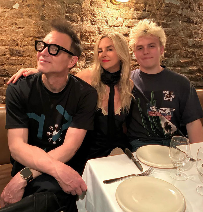 Mark Hoppus from Blink-182 sitting at a dining table with two others, brick wall background.