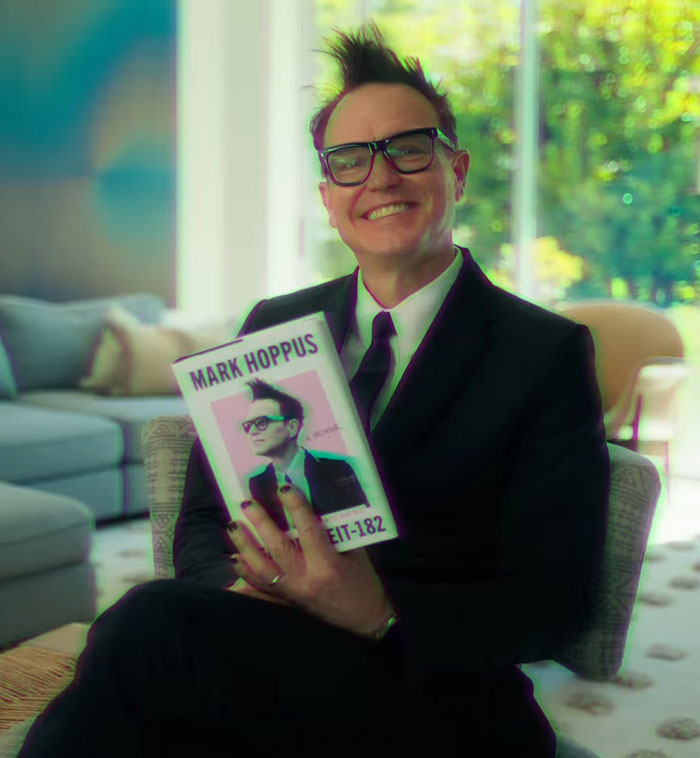 Mark Hoppus, seated in a living room, holds his Blink-182 book, smiling in a suit and glasses.