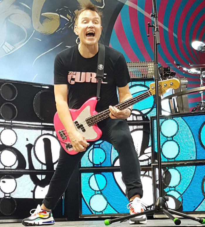 Mark Hoppus playing guitar on stage with a dynamic expression, wearing a "The Cure" shirt.