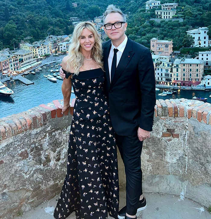 Mark Hoppus from Blink-182 in a suit with a woman in a dress, posing by a scenic waterfront view.