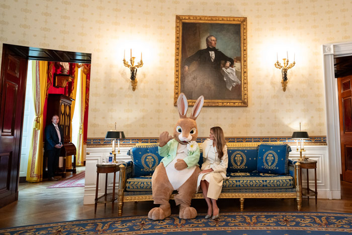 Melania Trump in White House interior with a person in a bunny costume, sitting on a blue and gold couch.