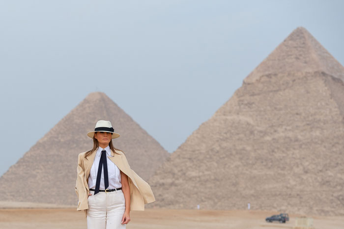 Woman in stylish outfit stands before pyramids, wearing a fedora. Glamorous setting evokes White House flair.