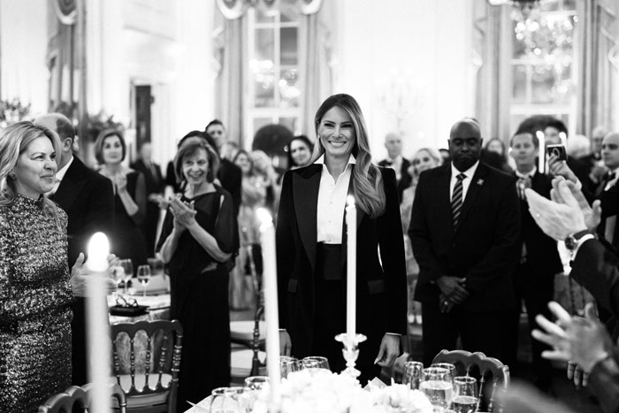 Melania Trump smiling in elegant attire at a White House event, surrounded by applauding guests.