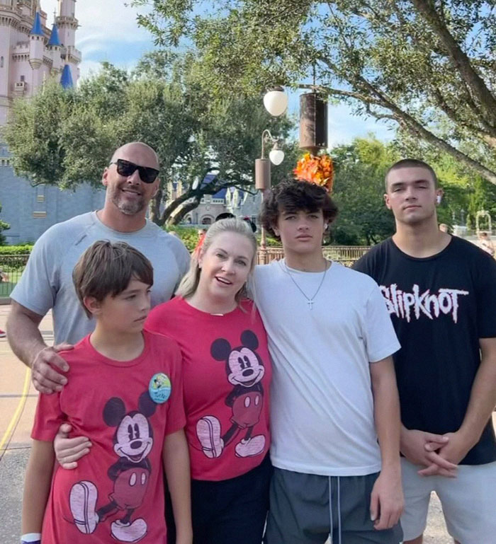 Family posing at a theme park, woman in a red Mickey shirt, castle backdrop, includes "Blink-182" and "Melissa Joan Hart".
