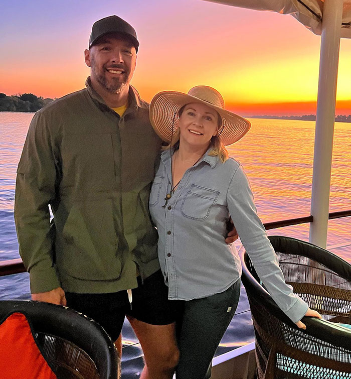 Couple smiling on a boat at sunset, with a beautiful orange sky in the background.