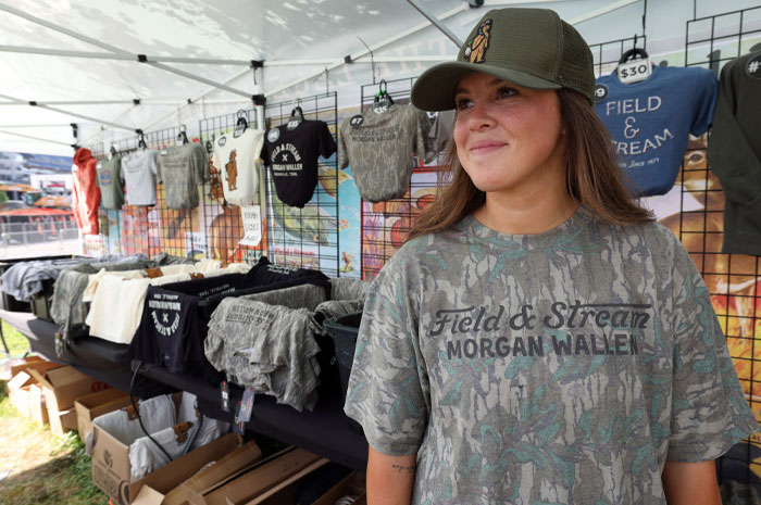 A woman in a "Morgan Wallen" camo shirt and cap at a merchandise stand at an outdoor event.
