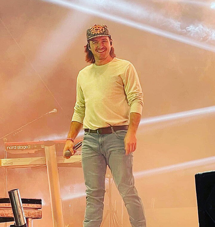 Man on stage holding microphone, wearing a casual outfit, with stage lights behind him.