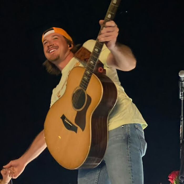 A smiling man in a backwards cap holds a guitar on stage, reaching out to the crowd.