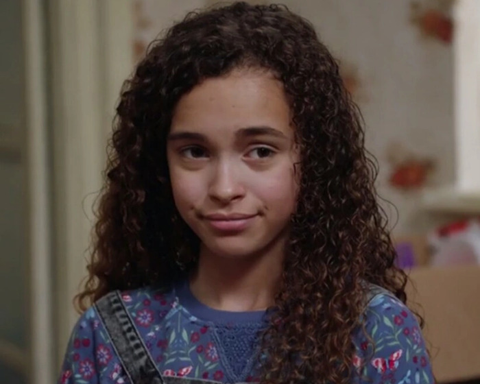 A young child actor with curly hair in a patterned top, smiling softly.