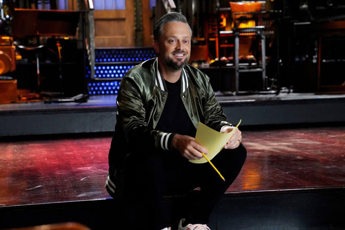 Nate Bargatze holding papers on stage, wearing a green jacket, spotlight on Nate Bargatze's net worth.