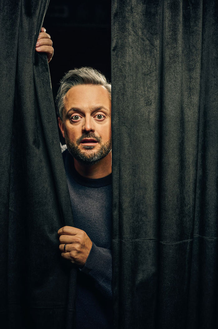 Comedian Nate Bargatze peeking from behind curtains, symbolizing success and net worth growth.