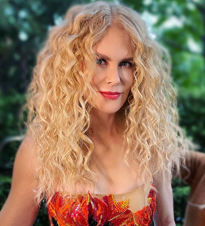 Nicole Kidman with curly blonde hair and red lipstick posing outdoors in colorful floral dress.